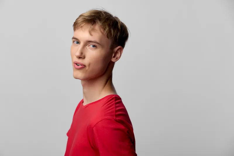 Man in a red shirt faces the camera with a friendly, playful expression in a neutral studio setting, conveying approachability, conversation, and confident communication.