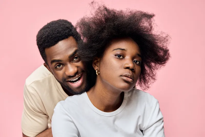 A joyful couple poses together in front of a pink background, showcasing natural hairstyles, vibrant emotions, and modern fashion choices, reflecting love and happiness