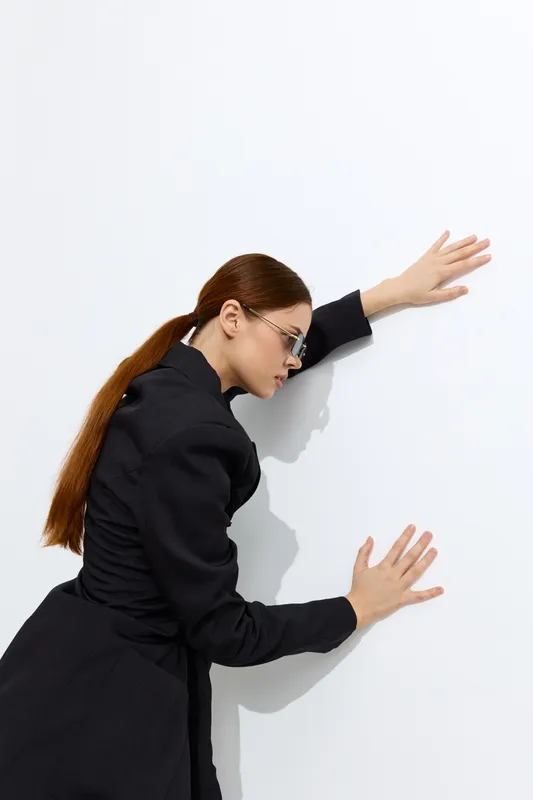 Fashion woman with long red hair and glasses wearing a black outfit leaning on a white wall, stylish and modern look with minimalistic background.