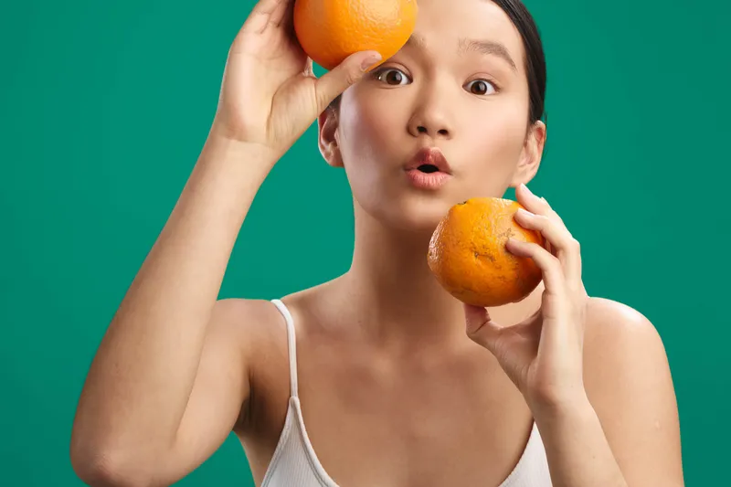 Cheerful asian woman holds bright oranges against a vivid green backdrop, smiling warmly and radiating a fresh, sunny vibe that feels inviting, playful, and stylish in a casual studio setting.