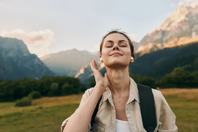 Cheerful woman outdoors, sunlit moment, relaxed pose, mountains in background, natural colors, backpack ready, fresh air, carefree mood, lifestyle vibe