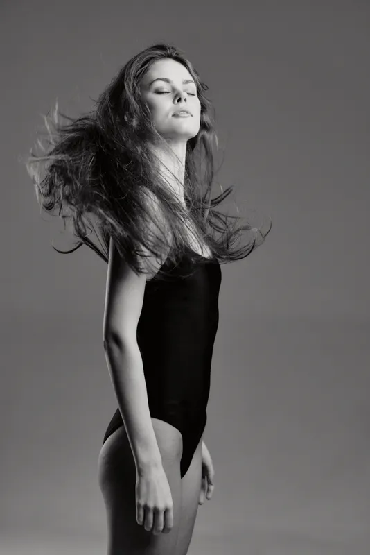 Black and white portrait of a young woman with long hair in a black bodysuit, expressing elegance and motion.