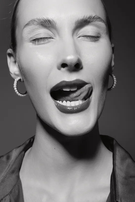 Close-up portrait of a fair-skinned white woman with closed eyes licking lips, wearing hoop earrings. Studio lighting highlights her expressive facial features and mood.