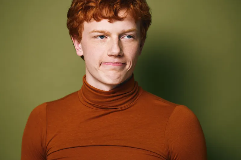 Young man with red hair and freckles wearing orange turtleneck sweater standing against green background with thoughtful expression. Portrait of casual style and natural light indoors.