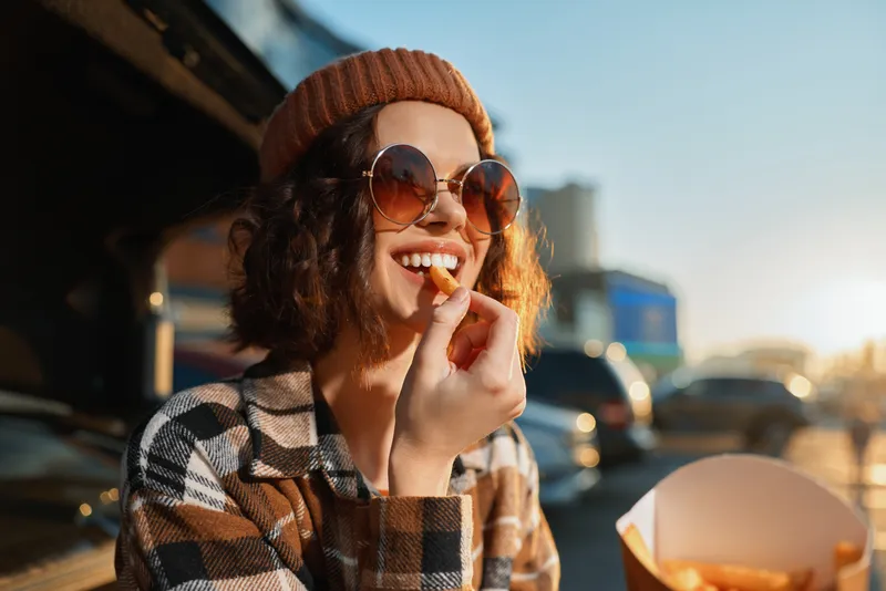 Woman smile sunglasses fries beanie golden hour urban authenticity candid lifestyle glow mindful living emotional storytelling. Young woman in plaid jacket eating fries outdoors at sunset, candid