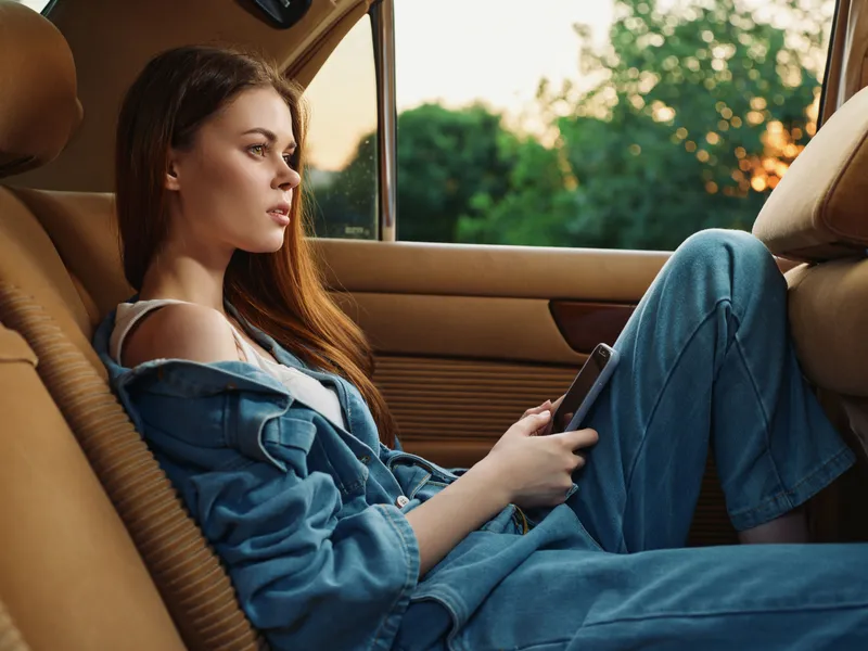 Young woman in denim outfit relaxing in a car while holding a phone, with a serene expression and lush greenery in the background during sunset.