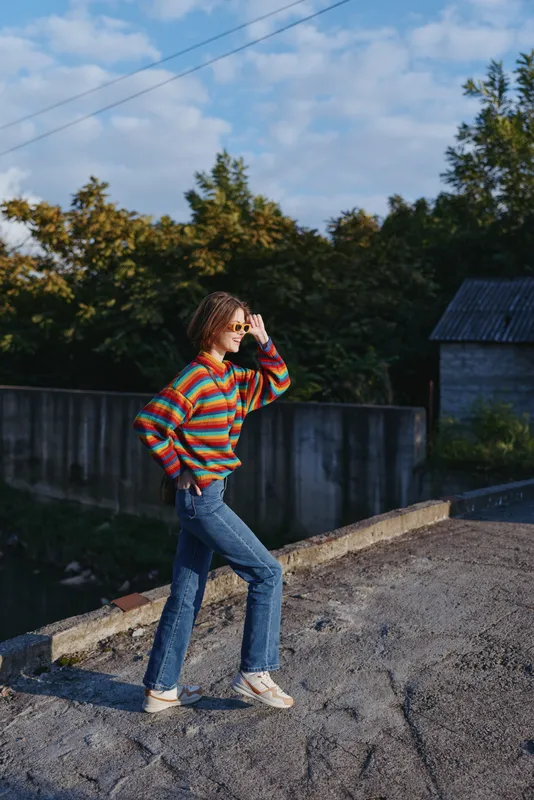 Woman in a colorful striped sweater and jeans outdoors, smiling and wearing sunglasses. Young woman in mid twenties posing on a sunny suburban road for travel lifestyle.