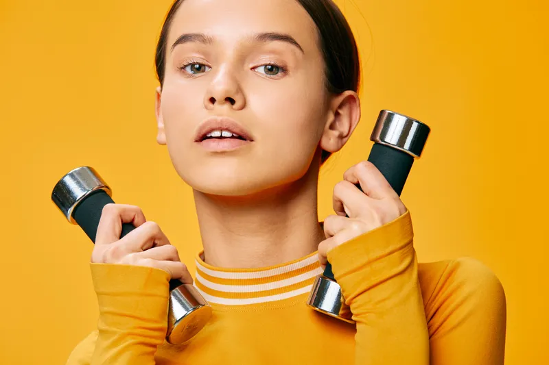 Female athlete in a bright yellow backdrop lifts dumbbells, showcasing a focused fitness routine with strong arms and core engagement. Dynamic sport scene emphasizing workout, training, and