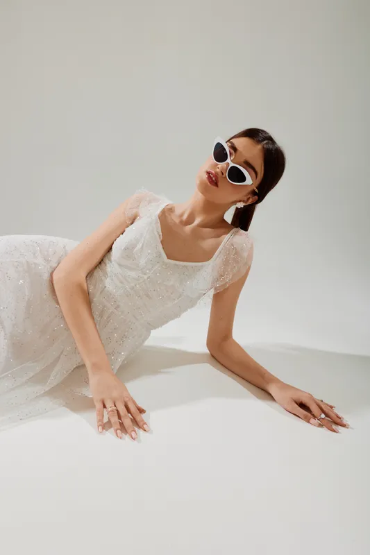 Young woman in a white elegant dress and stylish sunglasses lies on a white surface. Minimalist studio setting with soft lighting. Concept of beauty, fashion, and modern style.