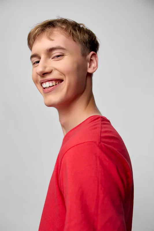 Smiling young man in a red shirt with casual attire, posed against a neutral backdrop, conveying warmth, approachability and positive energy for lifestyle and fashion projects
