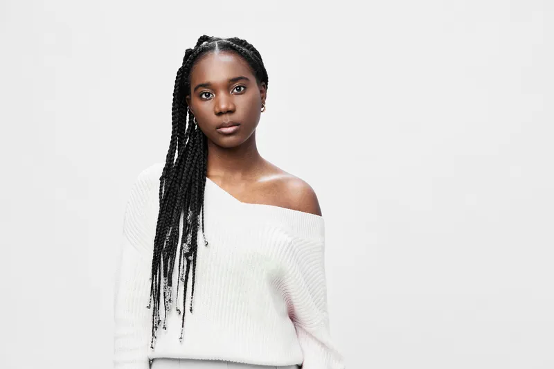 Portrait of a young black woman in a cozy white sweater, showcasing confidence and strength against a minimalistic light grey background