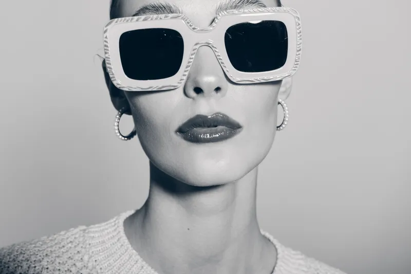 Close-up portrait of a fair skinned white woman wearing large square sunglasses and hoop earrings with glossy lips in monochrome studio lighting.