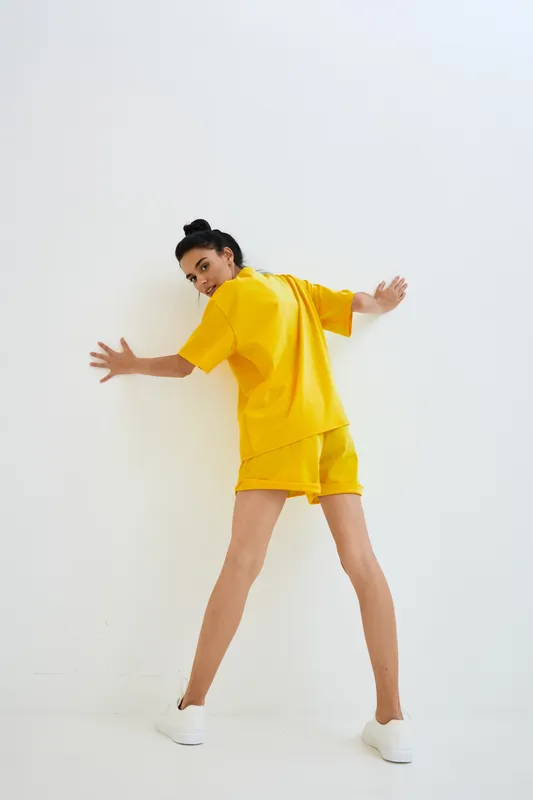 Young woman wearing bright yellow casual outfit and white sneakers posing against white wall, showing playful and dynamic body language with minimalistic background.