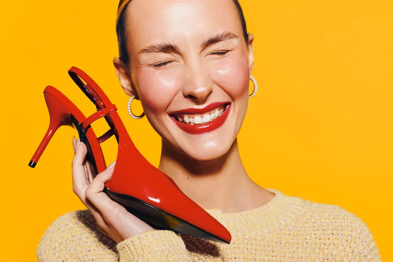Happy woman with closed eyes holds red high heel shoe near face on yellow background. Concept of fashion, style, beauty, and joyful emotions in studio lighting.