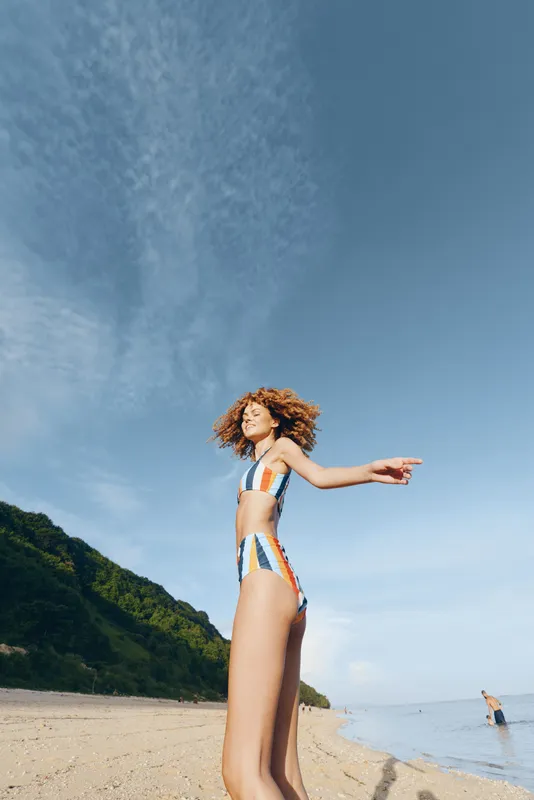 Young woman in striped swimsuit standing on sandy beach with green hills behind, pointing to the side, clear blue sky above, summer day, relaxed mood