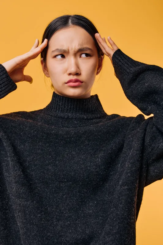Woman holding temples with fingers suffering from headache or stress, wearing black sweater against yellow background, showing discomfort and tension in face expression.