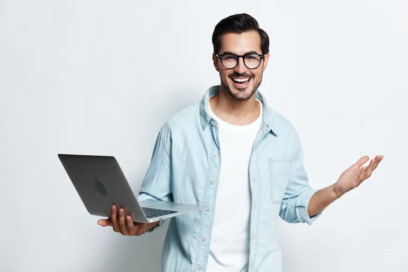 Cheerful smiling young man hold laptop, raise hand look at camera with excitement, dressed in casual blue shirt on light gray background. Tech lifestyle concept