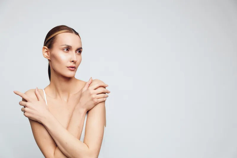 Young woman with clean skin and natural makeup poses with crossed arms and looks to side isolated on white background demonstrating beauty and skincare concept.