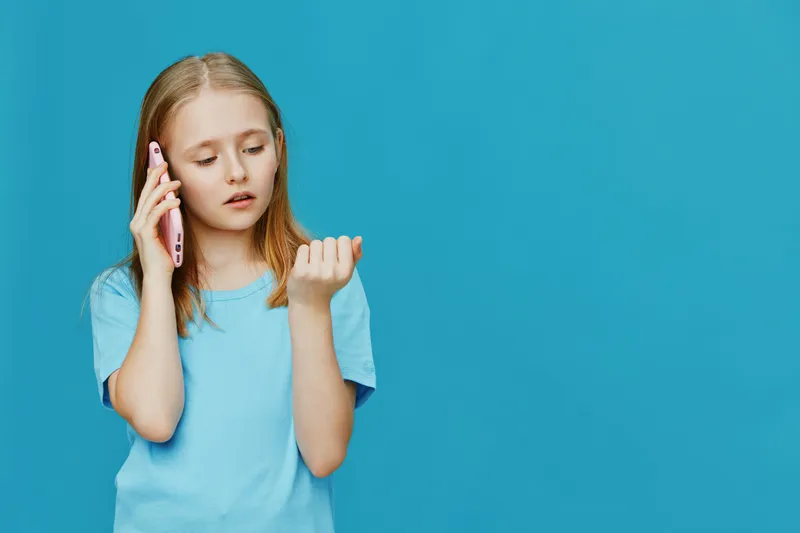 Young girl with long hair talking on a pink smartphone, wearing a light blue t shirt, showing curiosity against a bright turquoise background
