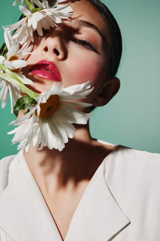 Close-up of a stylish woman with bold red lips holding large white daisies on her face. Concept for beauty, fashion, wellness, and artistic creativity.
