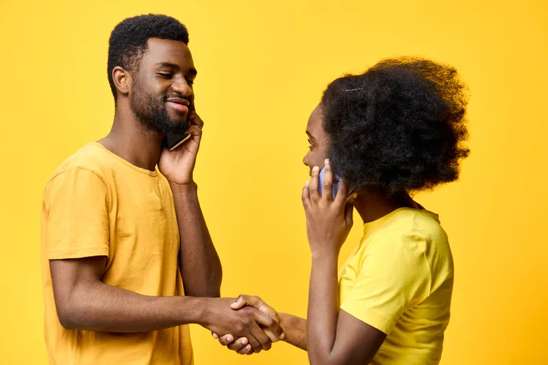 Young African couple communicating on mobile phones while shaking hands against a vibrant yellow background, showcasing connection and positivity