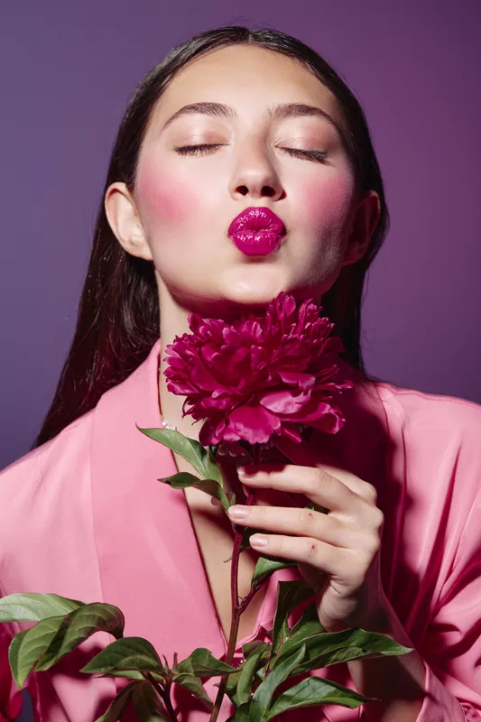 Young woman in pink attire blows a kiss with glossy fuchsia lips while holding a dark pink flower. Pastel purple background enhances the beauty creative fashion concept featured.