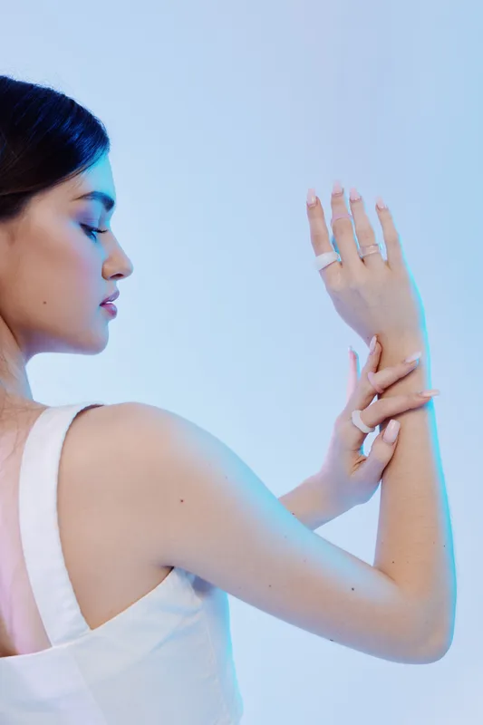 Young woman in white dress poses with closed eyes and raised arms against blue background. Soft lighting highlights her smooth skin and elegant posture. Concept of beauty, fashion, and grace.