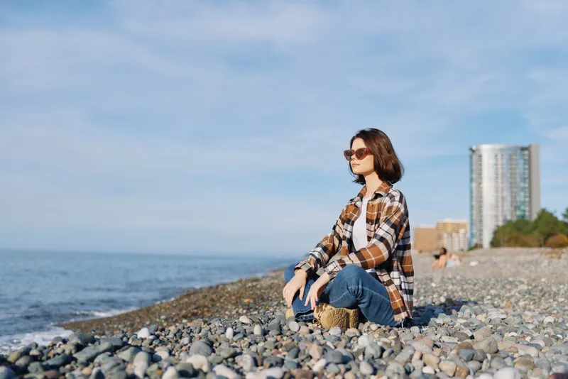 Beach, sea, woman sitting on pebbles by the shore, sunglasses, plaid shirt, casual outdoor relaxation in a sunny coastal moment