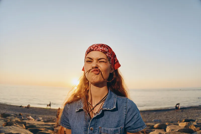 lifestyle woman in modern boho-western denim street style makes playful face with film color and warm sunset beach background atmosphere