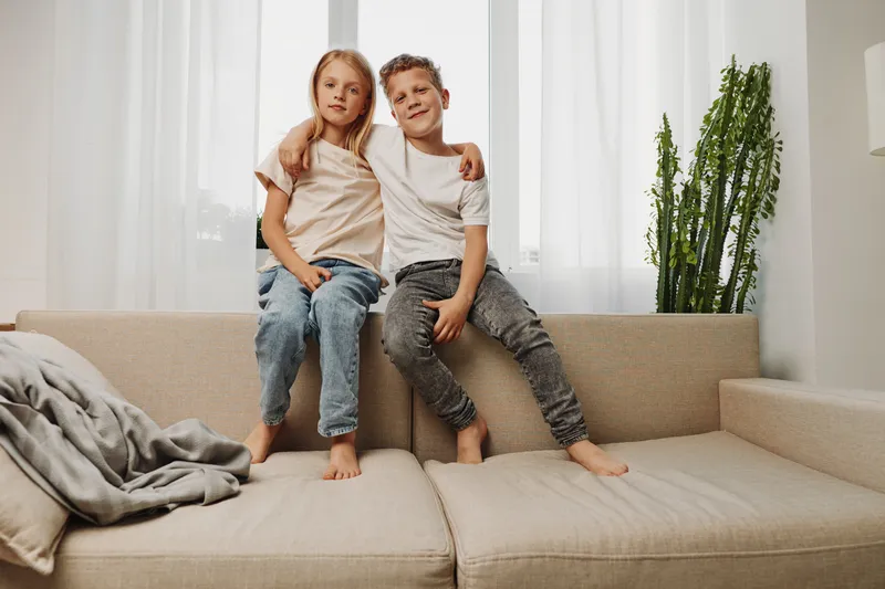 Two children sitting close together on a beige sofa in a bright living room, showing sibling bond and comfort. Casual clothes and relaxed barefoot pose create a warm home atmosphere.