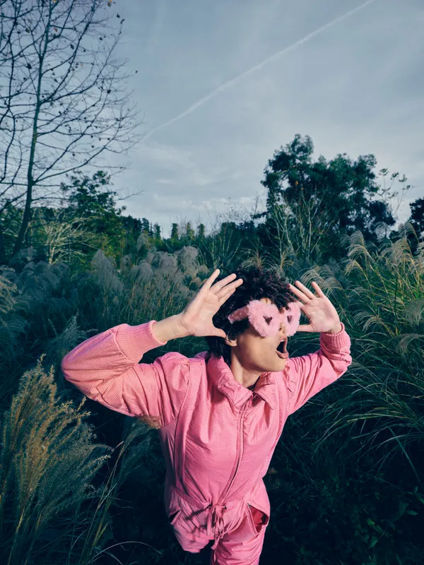 Outdoor portrait of a playful person in a pink jacket, surrounded by greenery and grass, capturing a casual selfie moment with cheerful expression in a natural setting.
