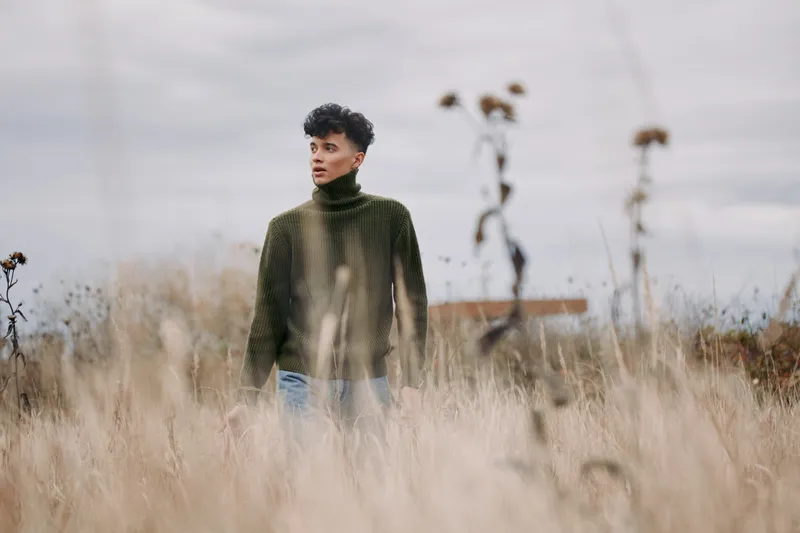 Young man in a rugged field wearing a green knit sweater, captured in a candid portrait conveying authenticity and a genuine, grounded presence amid tall dry grasses and overcast sky.