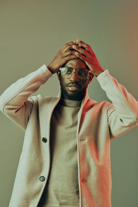A stylish young man expressing concern with his hands on his head, wearing a beige coat and sweater, set against a soft gradient background in warm tones