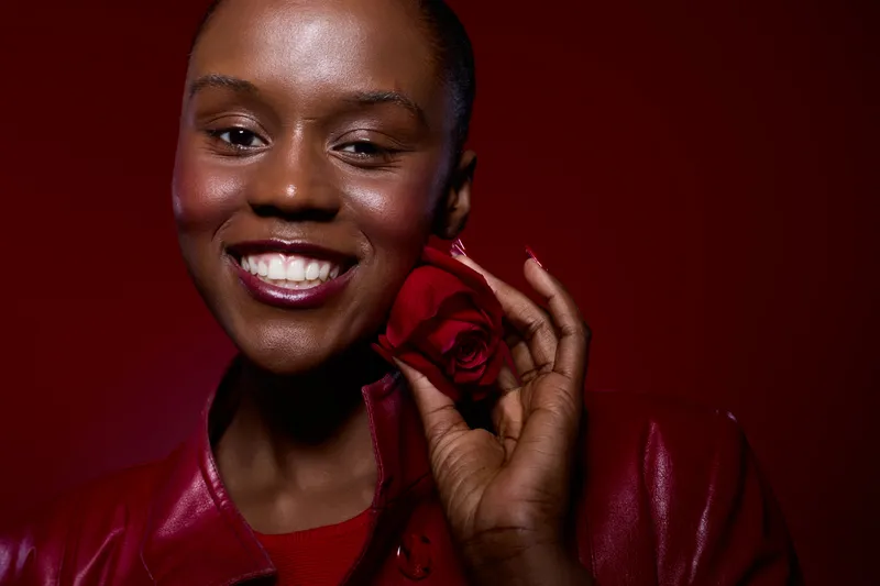 Close-up of a smiling African woman with dark skin holding a red rose near her face. She wears a red leather jacket against a dark red background. Concept of beauty, confidence, and elegance.