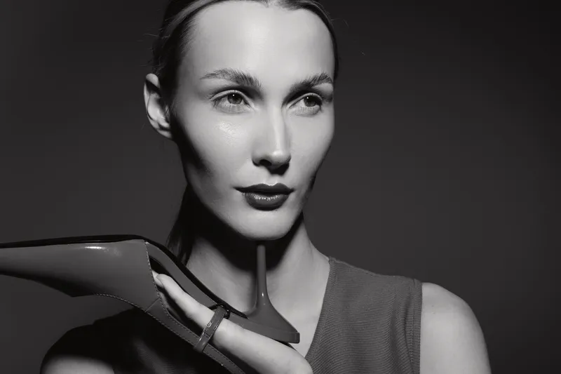 White woman with pale skin holds shoe near face in black and white studio portrait. Concept for fashion, elegance, beauty, and style with dramatic lighting and vintage mood.