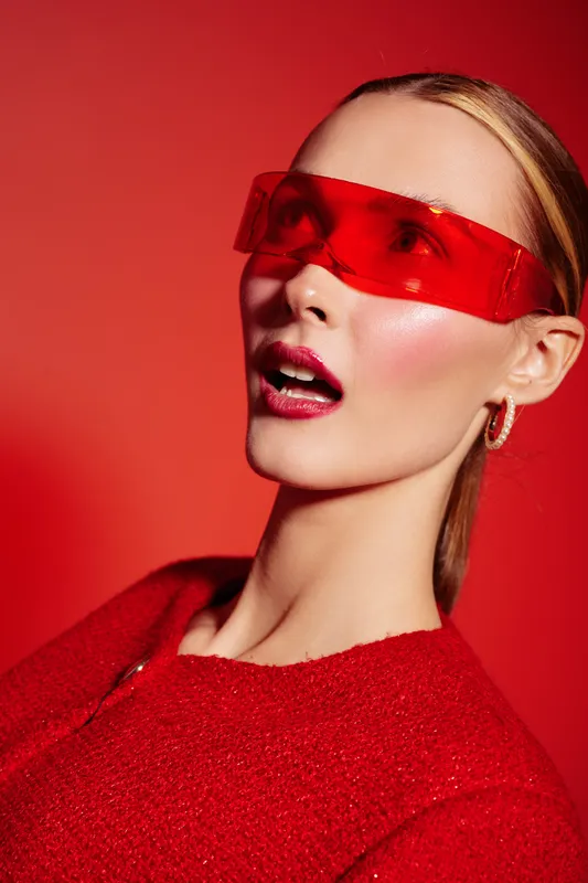 Portrait of a stylish woman wearing red futuristic sunglasses and a red textured sweater against a red background. Concept of fashion, modern style, and bold color contrast.