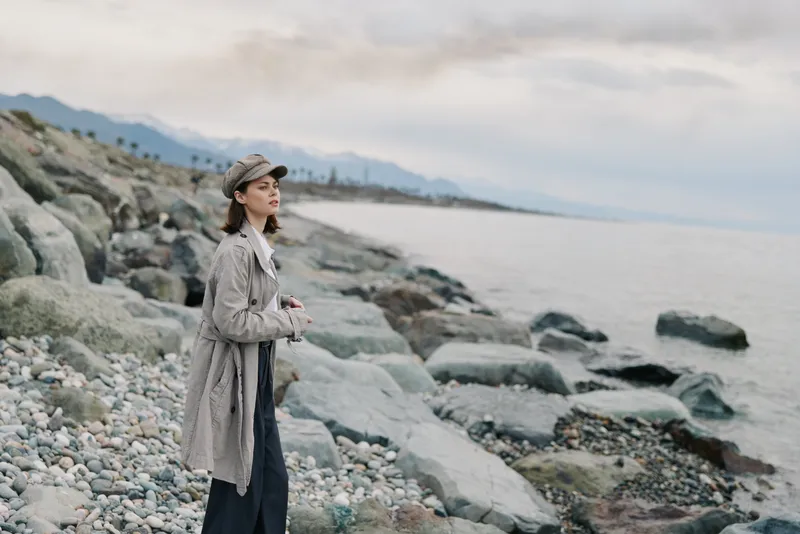 Young woman in casual coat and cap stands on rocky beach by the sea, looking into distance. Cloudy sky and mountains create calm natural landscape mood. Concept of travel, exploration, and solitude.