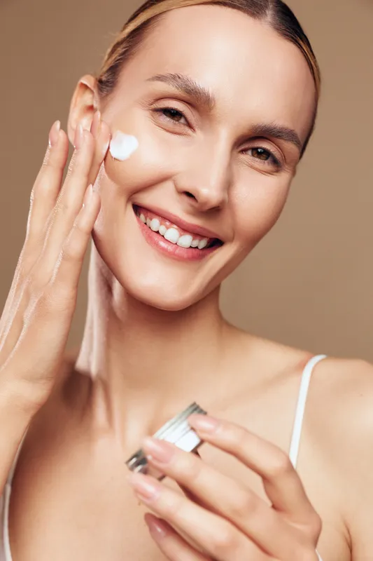 Smiling woman applies moisturizer cream on face with fingers holding jar skincare beauty routine natural skin care isolated on beige background closeup portrait.