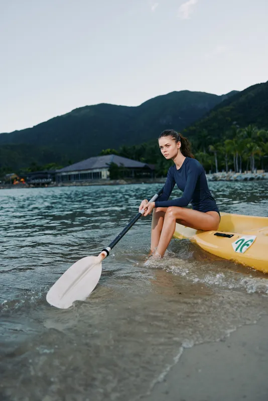 A female kayaker sits on a yellow kayak at the shoreline, paddle resting in the water, tranquil bay, distant mountains, clear sky, inviting calm adventure.
