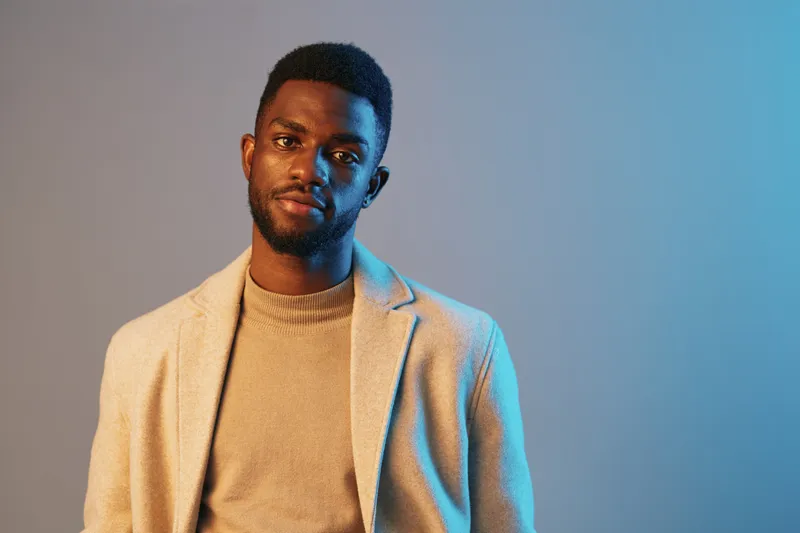 Stylish young African man in a beige blazer over a turtleneck sweater, exuding confidence against a soft blue and gray backdrop, showcasing modern fashion and elegance