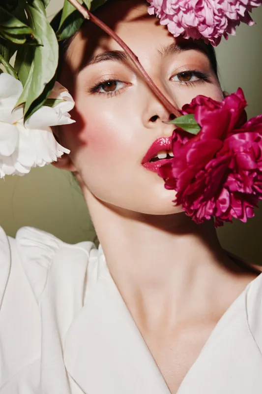 Young woman with fresh makeup featuring glossy lips and glowing skin posing creatively with large pink and white flowers on soft green background for beauty fashion concept.