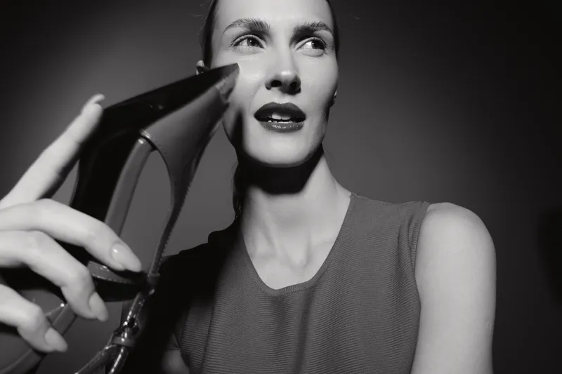 Elegant white woman with pale skin holds black high heel shoe near face in studio. Concept of fashion and style.