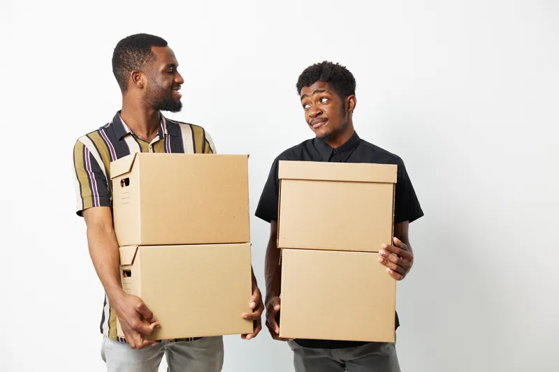 Two smiling black men are holding cardboard boxes, conveying a sense of teamwork and collaboration in a bright and minimalistic setting.