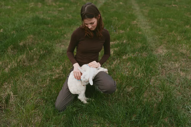 Young woman in brown sweater kneels on grass while gently holding a white baby goat outdoors in a natural lifestyle setting during daytime.