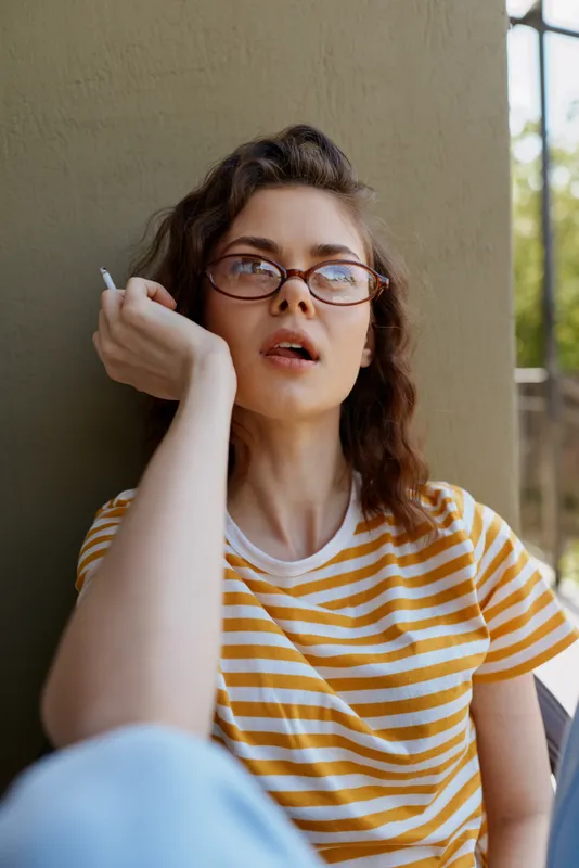 Casual woman at home, wearing a striped shirt and glasses, relaxed pose and thoughtful gaze, soft daylight, warm tones, cozy atmosphere, everyday lifestyle portrait