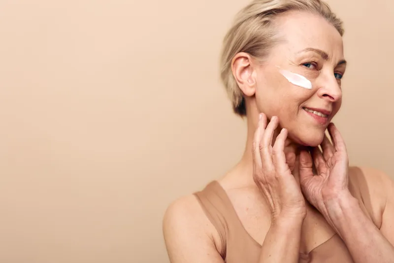 Smiling mature woman applies white face cream on cheek, touching skin gently. Nude background highlights skincare, beauty, antiaging, wellness, and natural selfcare concept.