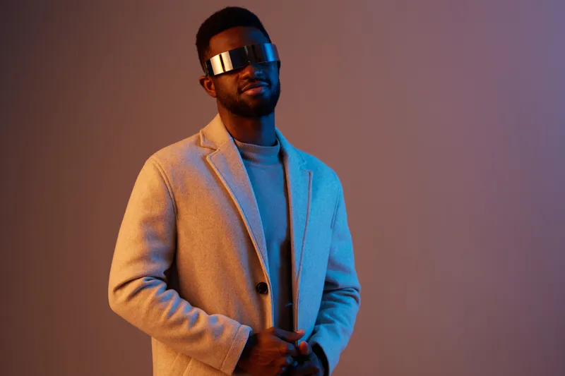 Stylish young man wearing futuristic glasses and a beige coat against a blue and purple background, exuding confidence and a modern vibe
