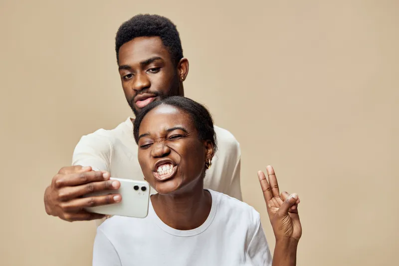 A joyful young Black couple taking a selfie against a beige background, showcasing playful expressions and a fun atmosphere, perfect for social media themes