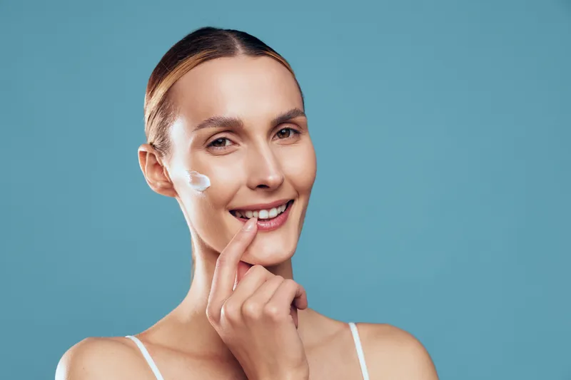 Smiling woman applying face cream, clear skin, natural beauty, skincare routine, blue background, soft lighting, closeup portrait, healthy lifestyle, wellness concept