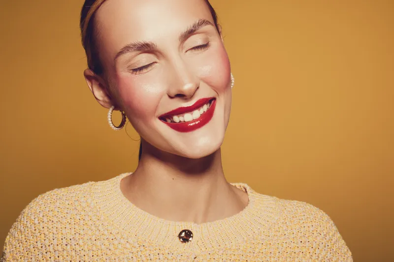 Smiling woman with closed eyes wearing red lipstick and yellow sweater poses against orange background. Concept of happiness, beauty, makeup, and fashion portrait with warm lighting.